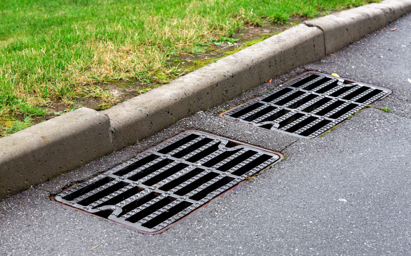 manhole cover rain grate on an asphalt road near a stone curb on the side of the road green grass.